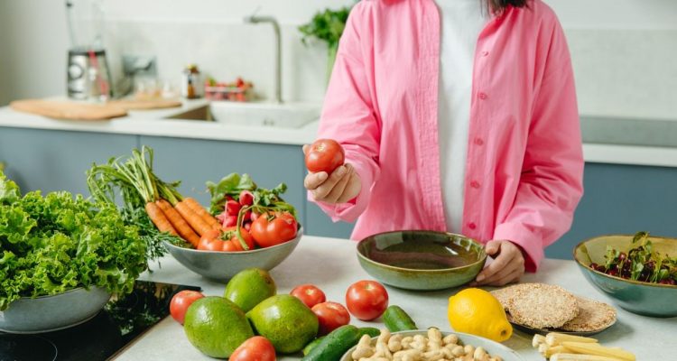 Woman in pink overshirt holds tomato in kitchen with fresh fruits and veggies.