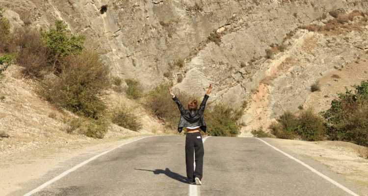 A woman in a leather jacket stands joyfully on a rural road surrounded by rocky terrain.